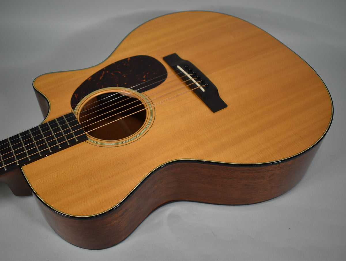 Close-up of a light-wood acoustic guitar, angled to show the body, soundhole with green trim, dark pickguard, and bridge.