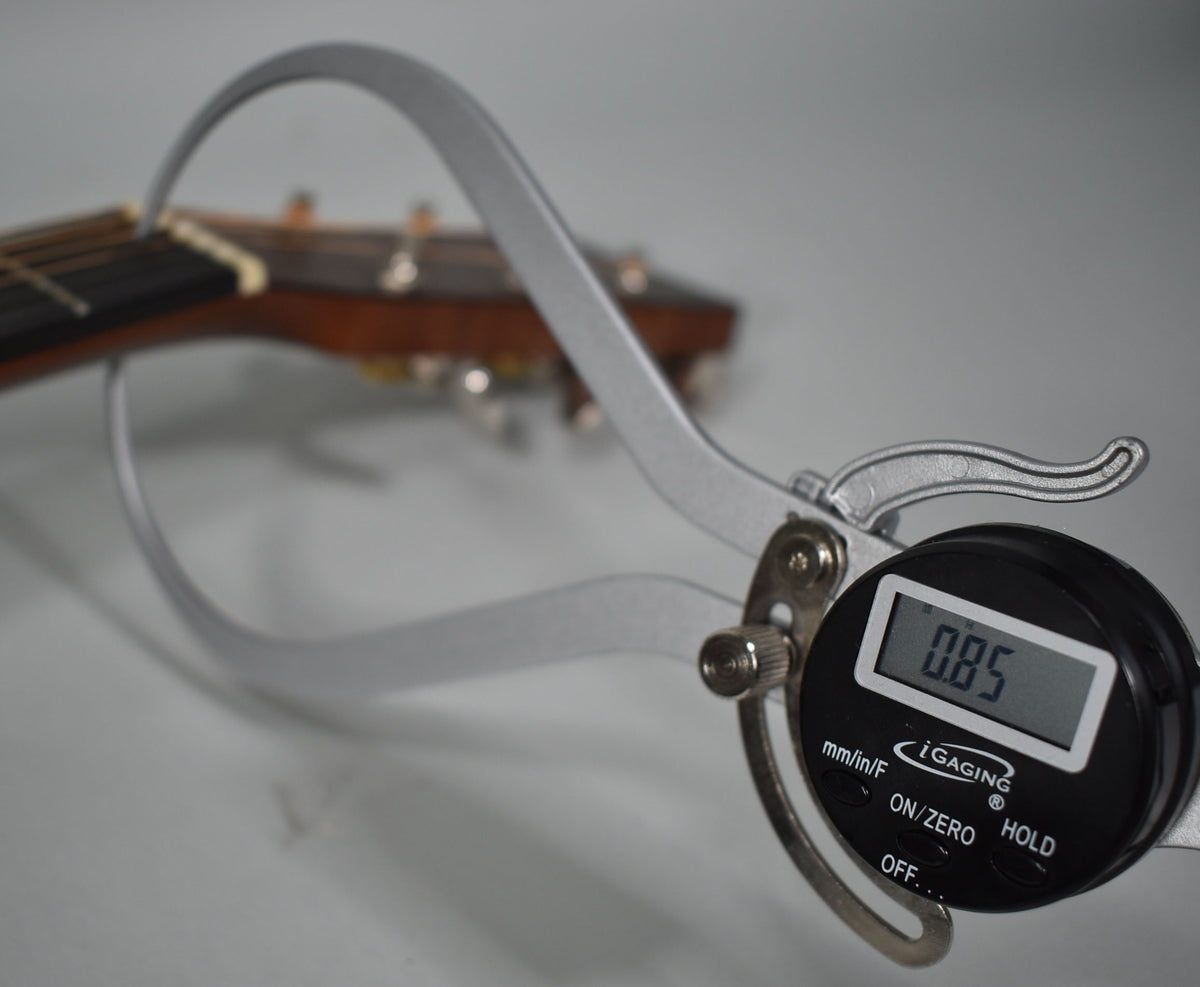 A digital caliper measures the guitar's nut, showing 0.85, with the headstock and tuning pegs blurred in the background.