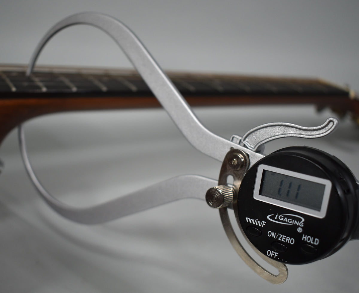 A digital caliper measures the guitar's fretboard, showing a reading of 1.11, with the neck and frets blurred in the background.
