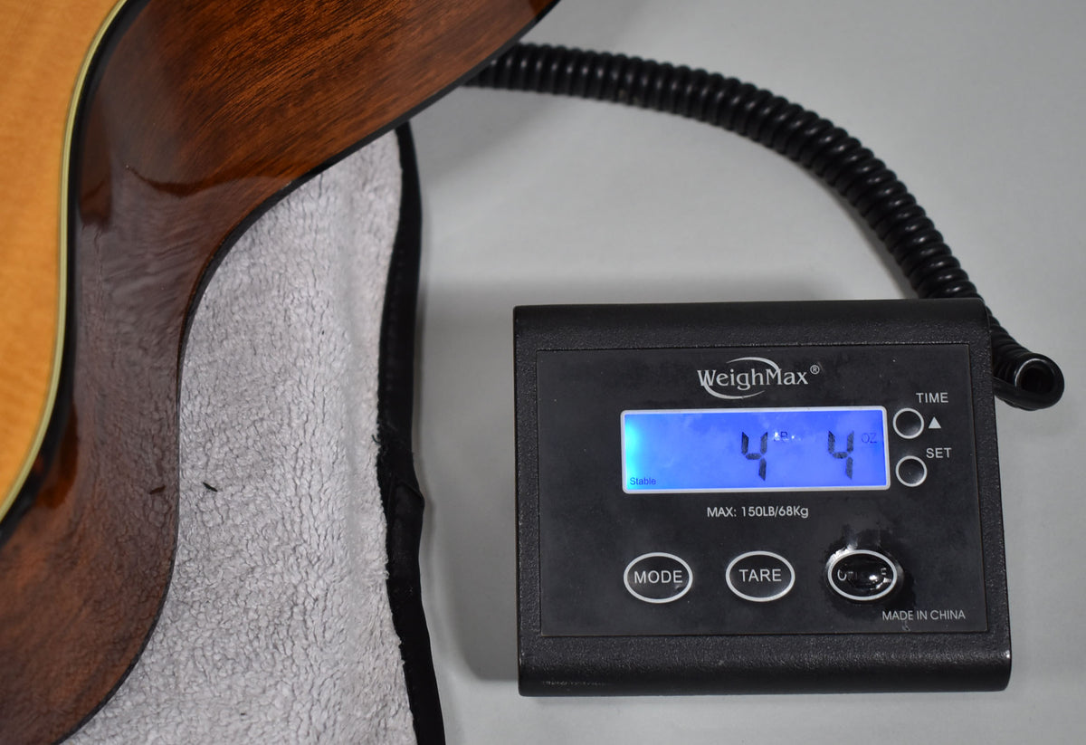 The guitar's dark brown back and side are visible, resting on a white towel next to a digital scale displaying "4 lbs 4 oz".