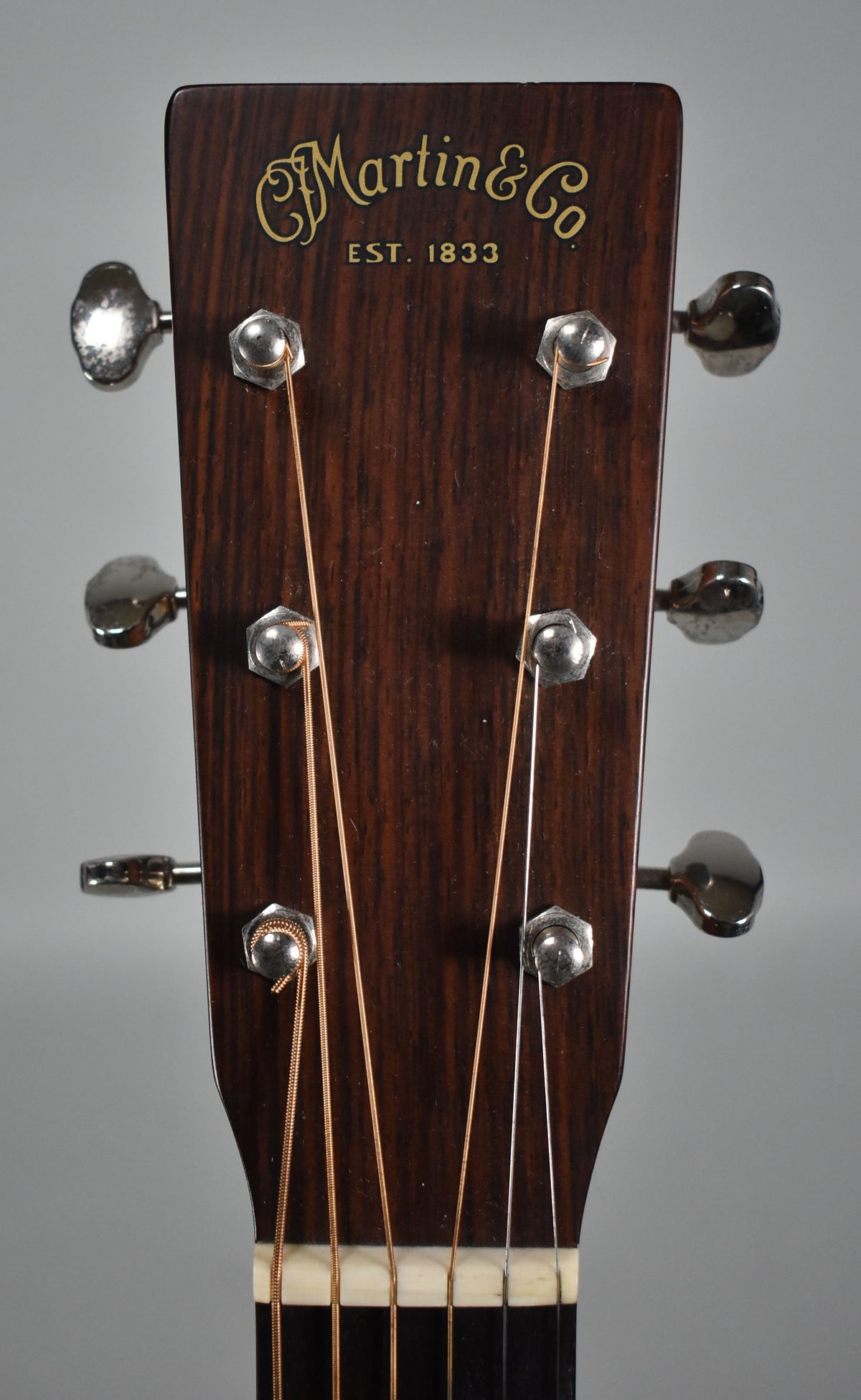 The dark brown headstock, viewed from the front, features six chrome tuners and a gold logo, with light and dark strings.