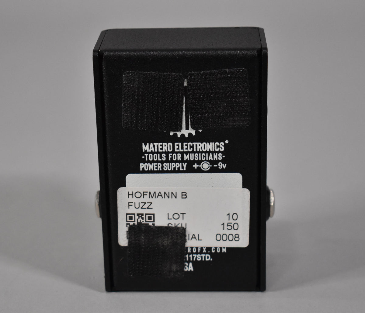Rear view of a matte black fuzz pedal, with two strips of black hook-and-loop tape and a partially obscured white label.