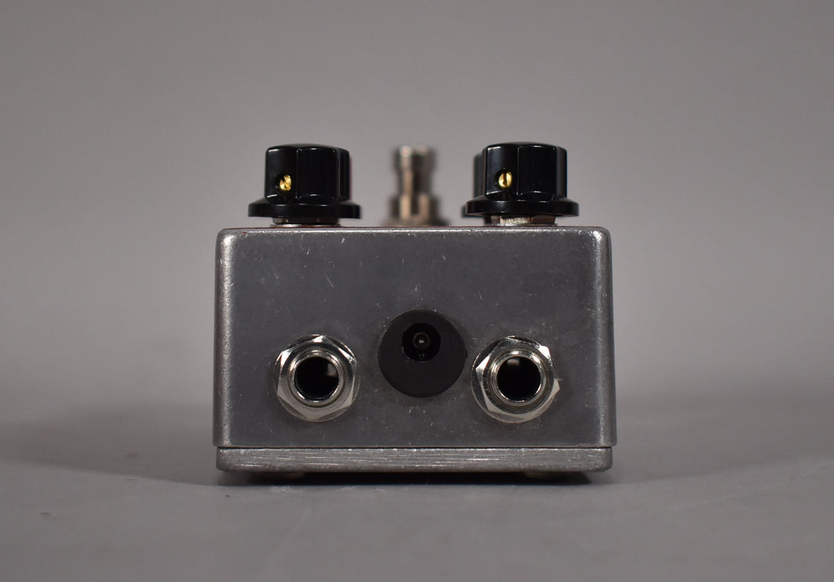 The silver pedal is viewed from the back, showing two black knobs on top, two input jacks, and a power input.