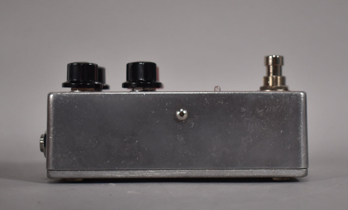 Side view of the brushed aluminum pedal, showing two black knobs, a footswitch, and an input jack on the left.