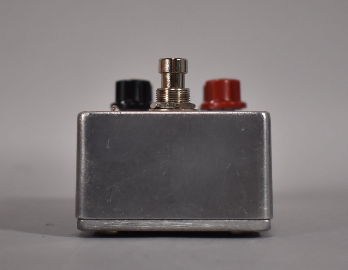 The brushed aluminum pedal is viewed from the front, showing a silver footswitch, a black knob, and a red knob.