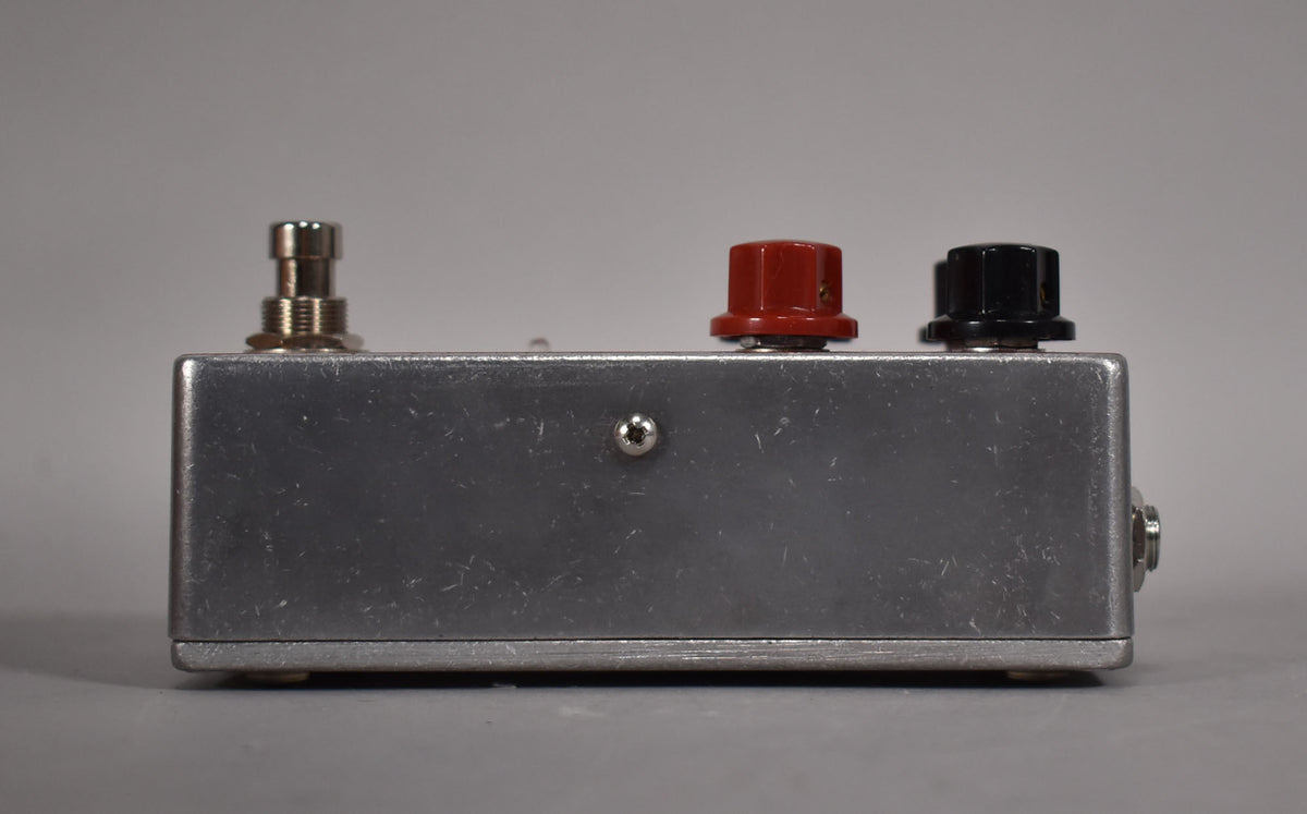 Side view of the brushed aluminum pedal, showing the footswitch, a red knob, a black knob, and an input jack on the right.