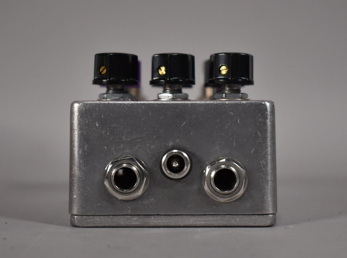 The silver-grey pedal is viewed from the back, showing three black knobs with gold indicators, two input jacks, and a power input.