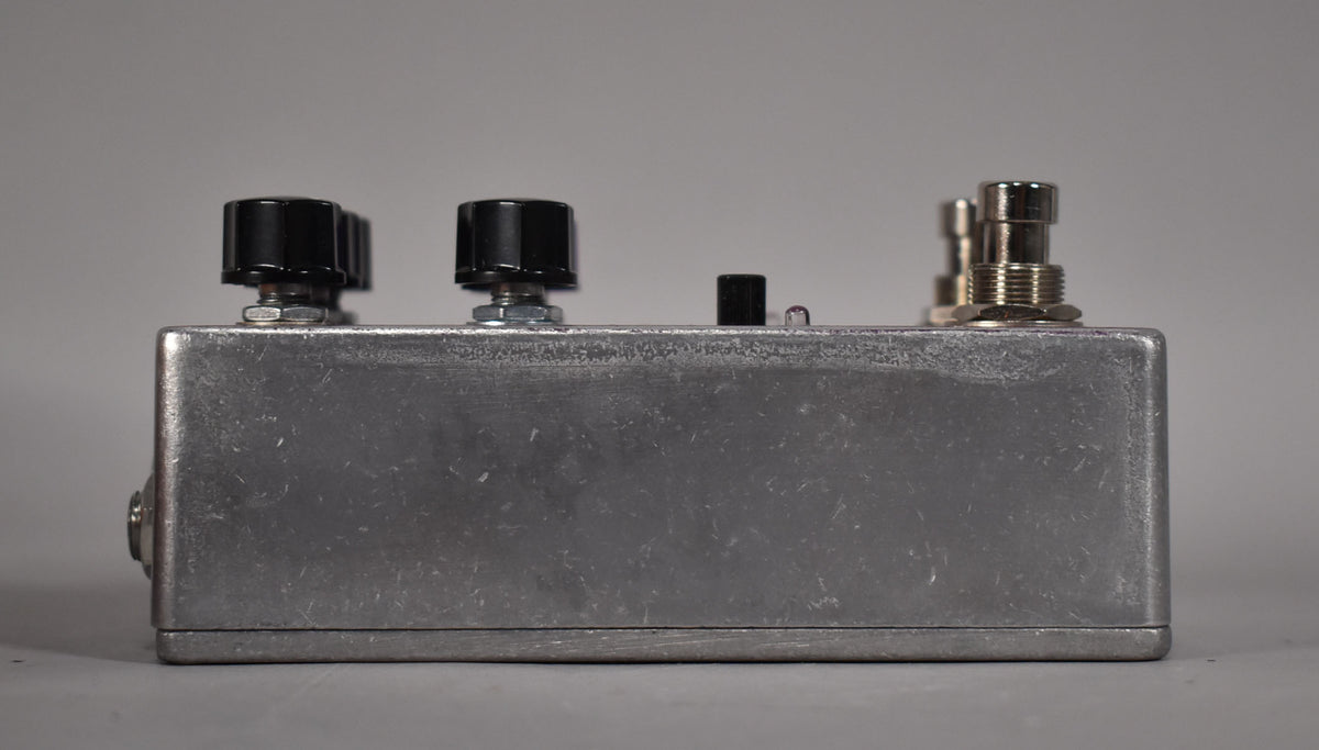 The brushed aluminum pedal is viewed from the side, showing two black knobs, a small black switch, and two silver jacks.