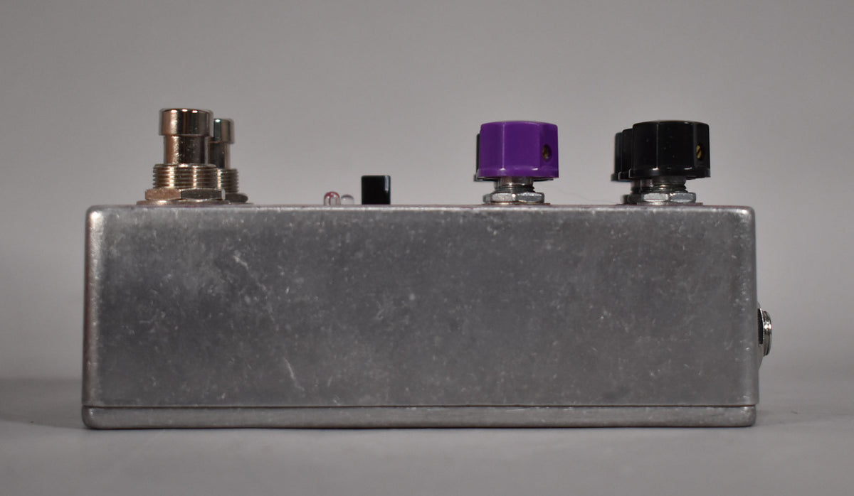 Side view of a brushed aluminum pedal with two silver footswitches, a small black switch, a purple knob, and a black knob.