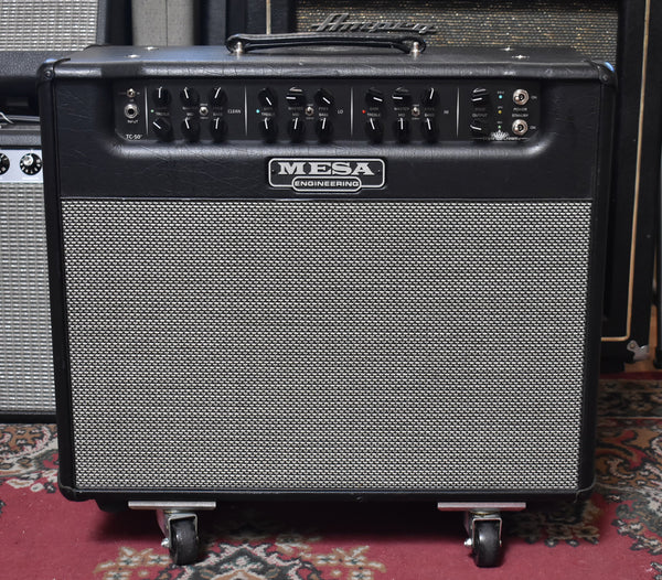 Front view of a black combo amp with a silver grille, black control panel with 13 knobs, and four casters on the bottom.