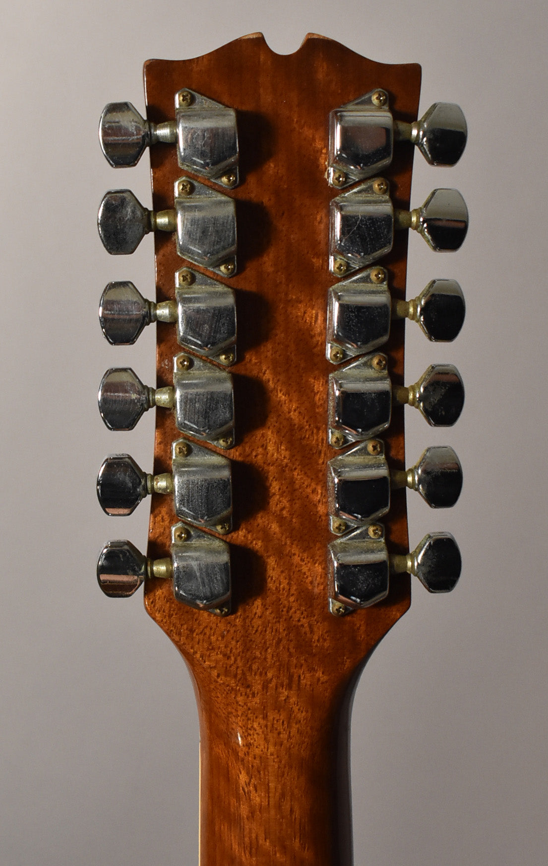 The back of a natural wood headstock and neck, showing 12 tarnished chrome tuners in two rows of six, with visible wear.