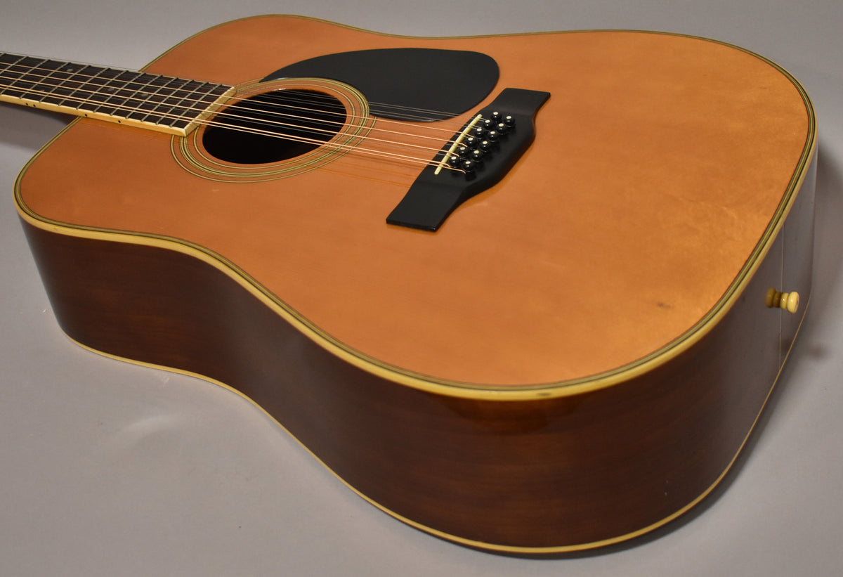 The natural finish 12-string dreadnought is angled from the upper left, showing the body, soundhole, black pickguard, and bridge.