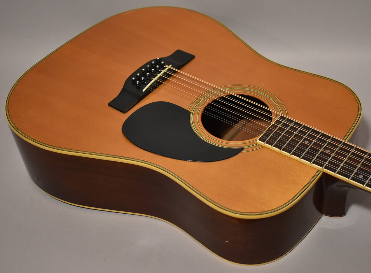 The natural finish 12-string dreadnought is angled from the upper left, showing the body, soundhole, black pickguard, and lower fretboard.