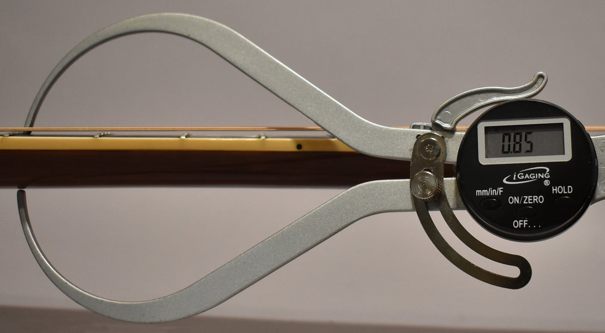 A digital caliper measures the guitar's natural-finish neck and fretboard, showing a reading of 0.85.