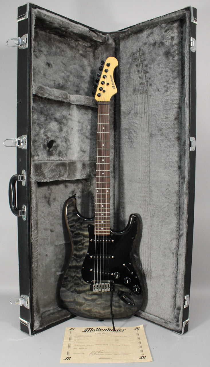 Full view of a gray flamed-top electric guitar with a black pickguard and controls, standing in an open, gray-lined hard case.
