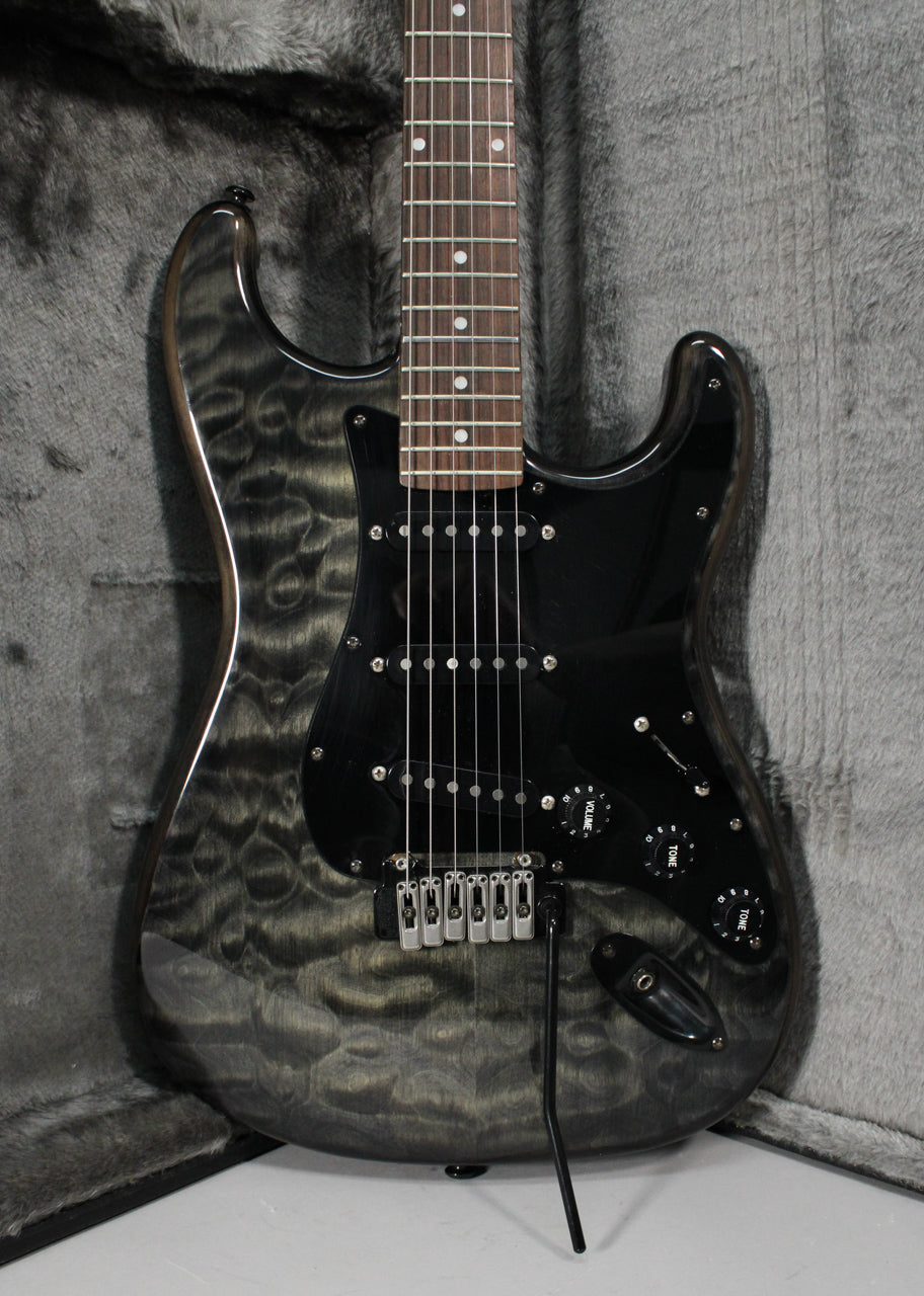 A gray-black electric guitar with a quilted maple top, black pickguard, and tremolo bar, resting in a gray plush case.