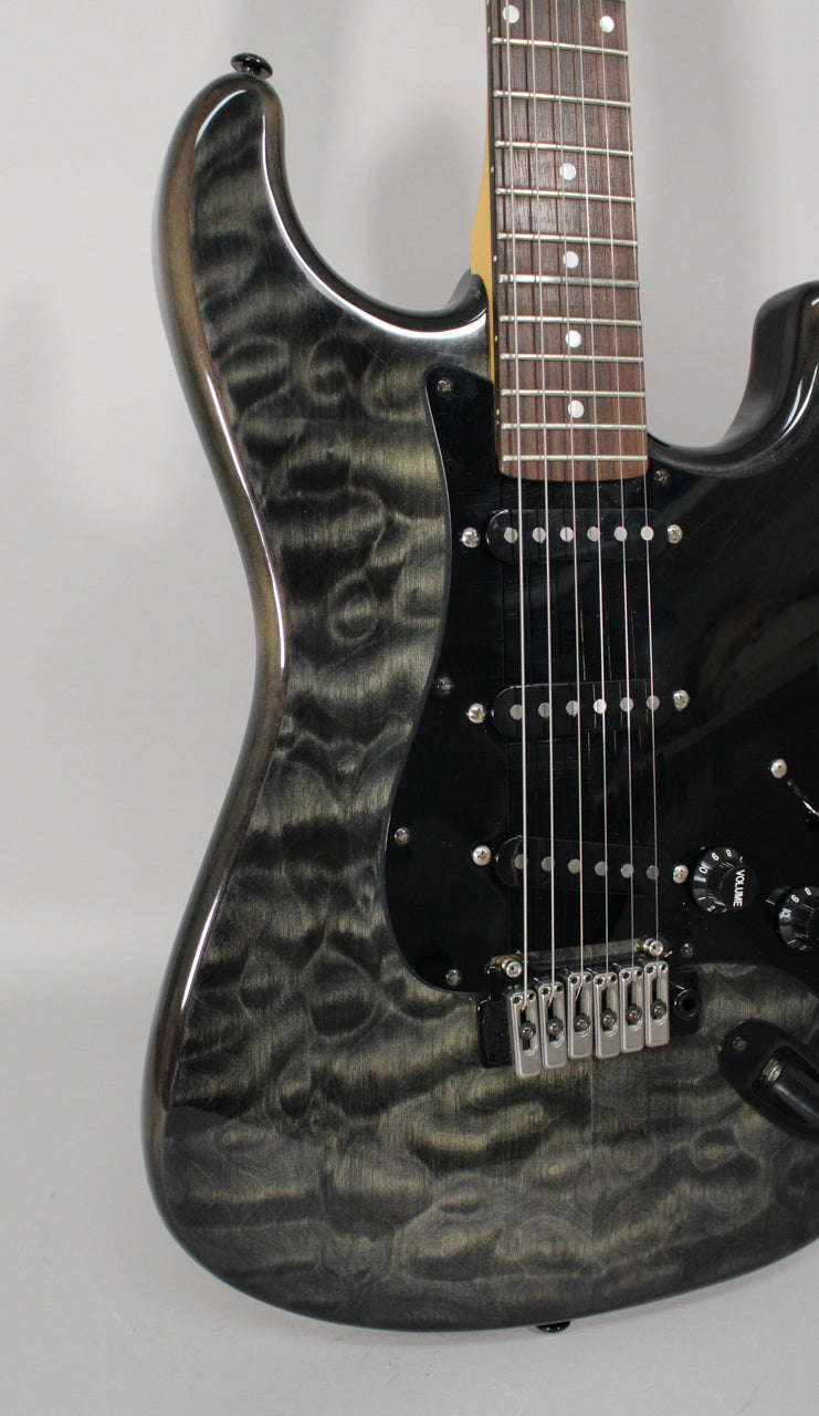Close-up of a gray electric guitar with a wavy wood grain, black pickguard, three single-coil pickups, and a tremolo bridge.