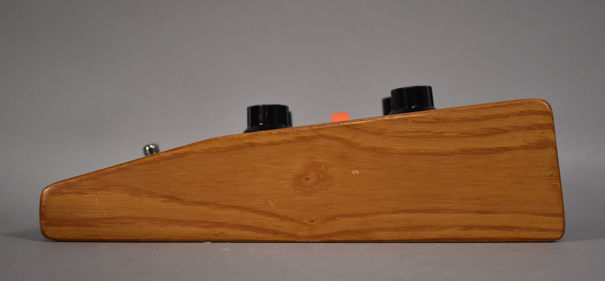 The right side of the pedal is shown, revealing its light brown wood casing with visible grain, two black knobs, and a small orange indicator.