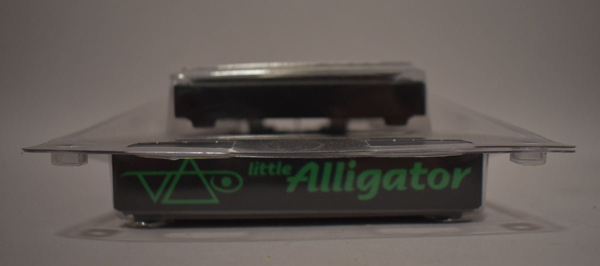The black pedal, still sealed in its clear plastic blister packaging, is viewed from the front, showing green lettering.