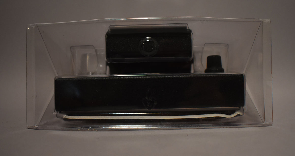 The black pedal is seen from the front, still sealed in its clear plastic blister packaging, with a small black knob on the right.