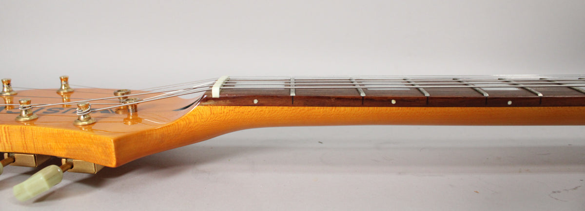 Side view of the headstock and neck, showing the light amber finish, gold tuners, white nut, and dark rosewood fretboard.