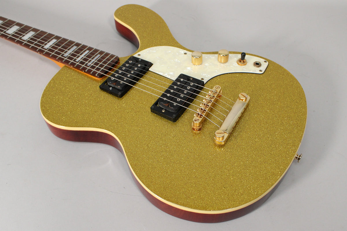 The gold sparkle guitar body, with a pearloid pickguard, two black humbuckers, and gold hardware, is angled from the upper left.