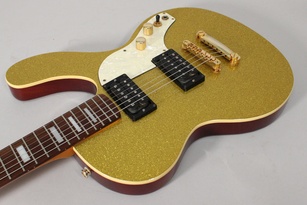 Angled view of the gold sparkle guitar body, showing two black humbuckers, gold hardware, and a white pearloid pickguard.