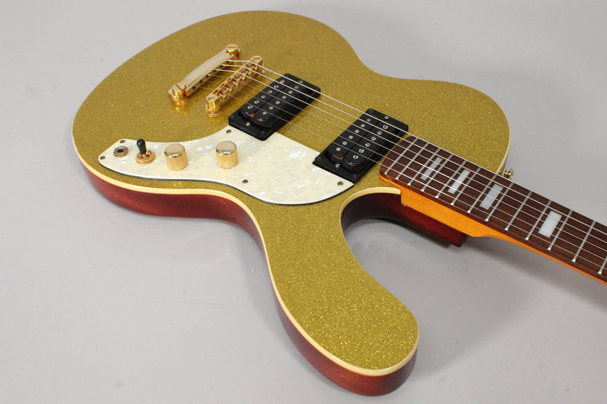 The gold sparkle guitar body is angled, showing two black humbuckers, a pearloid pickguard, and gold hardware.