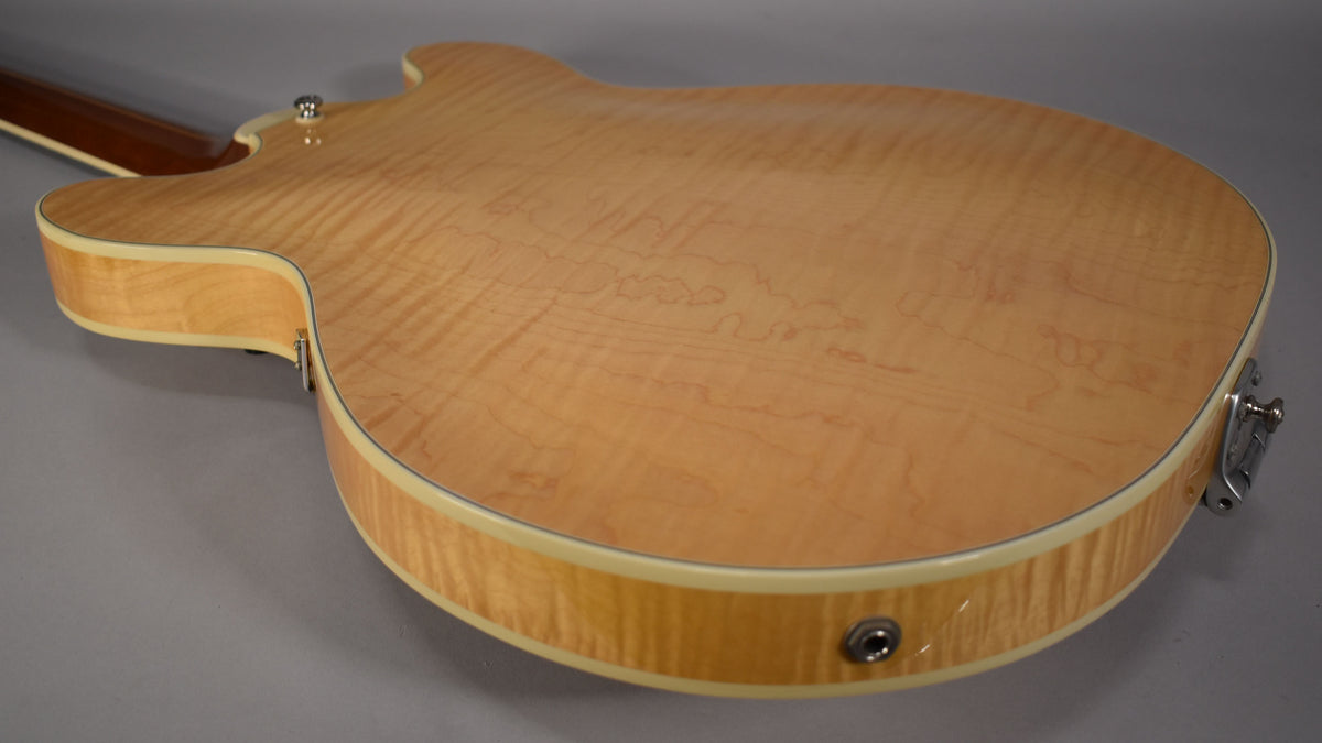 The back of a natural finish hollow-body guitar, showing flame maple grain, cream binding, and a chrome output jack and strap button.