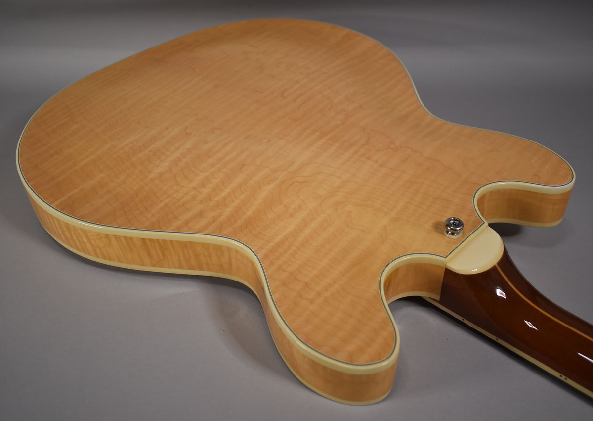 The back of the natural finish guitar shows flame maple grain, cream binding, and a chrome output jack near the neck heel.