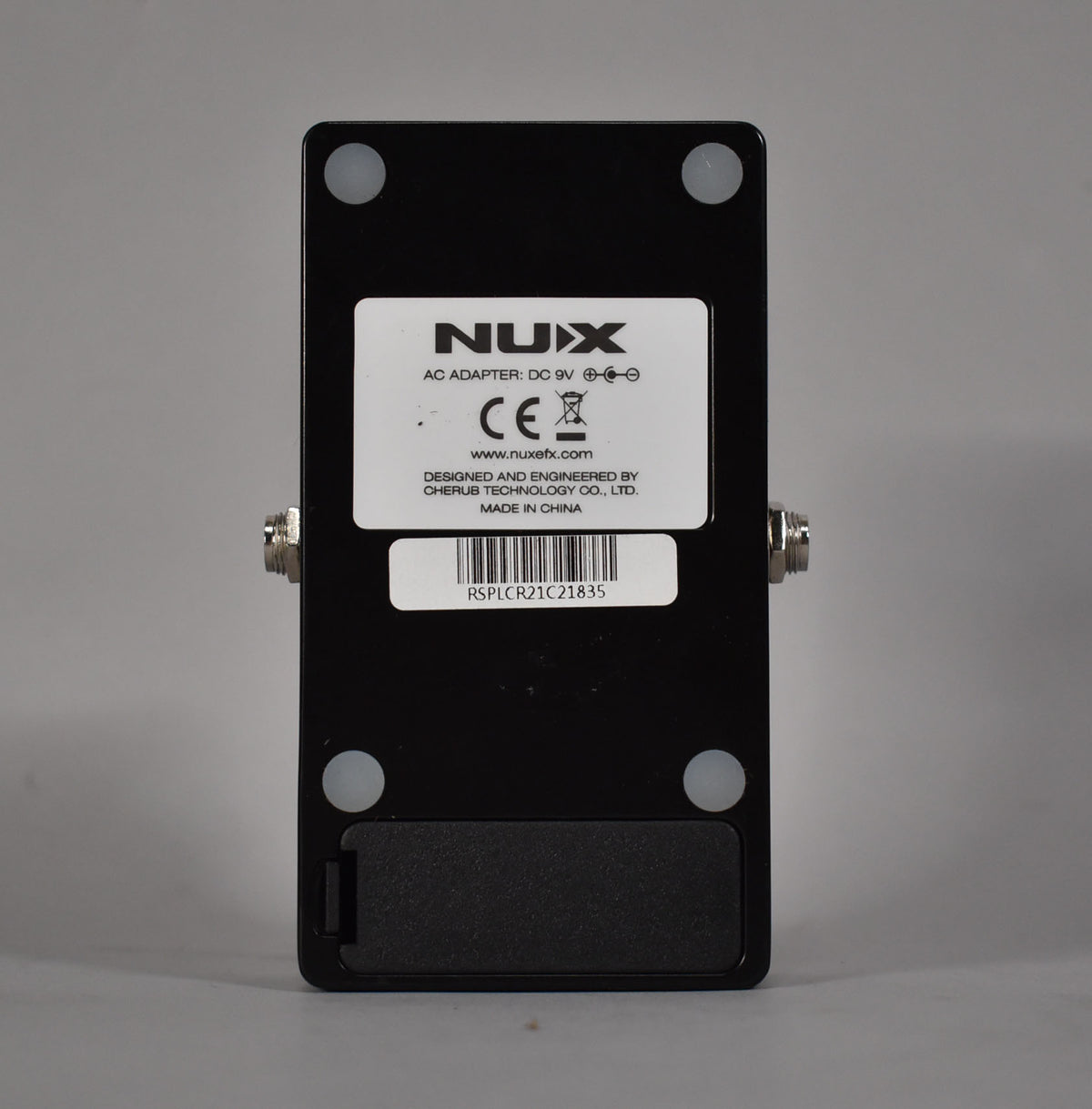 The pedal's black metal casing is viewed from the back, showing a white label, four rubber feet, and a battery compartment.