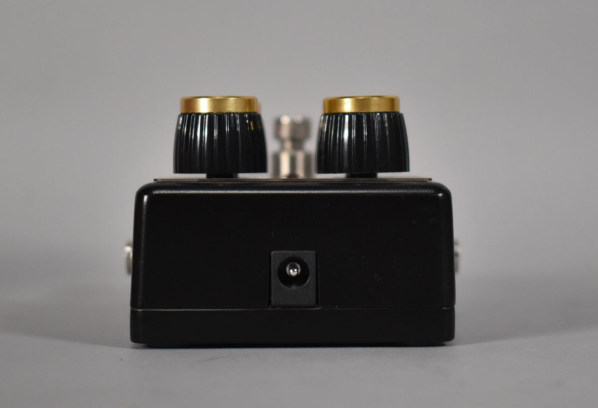 The black pedal is viewed from the rear, showing the power input jack and two black ridged knobs with gold tops.