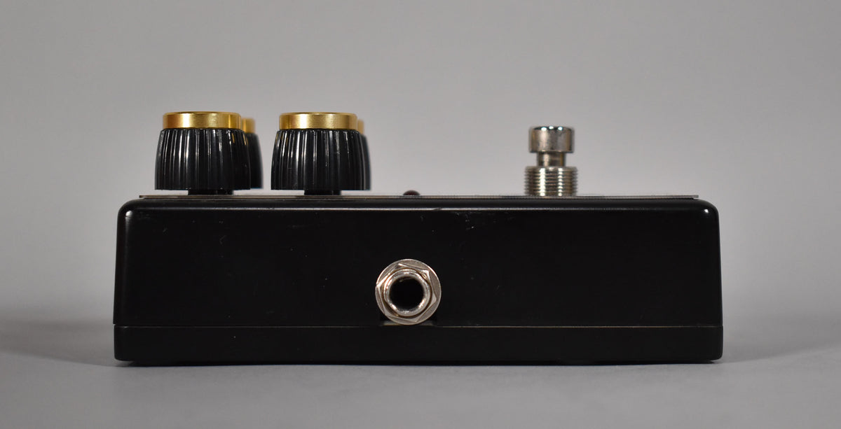 The black pedal is viewed from the side, showing a silver input jack, two black ridged knobs with gold tops, and a silver footswitch.