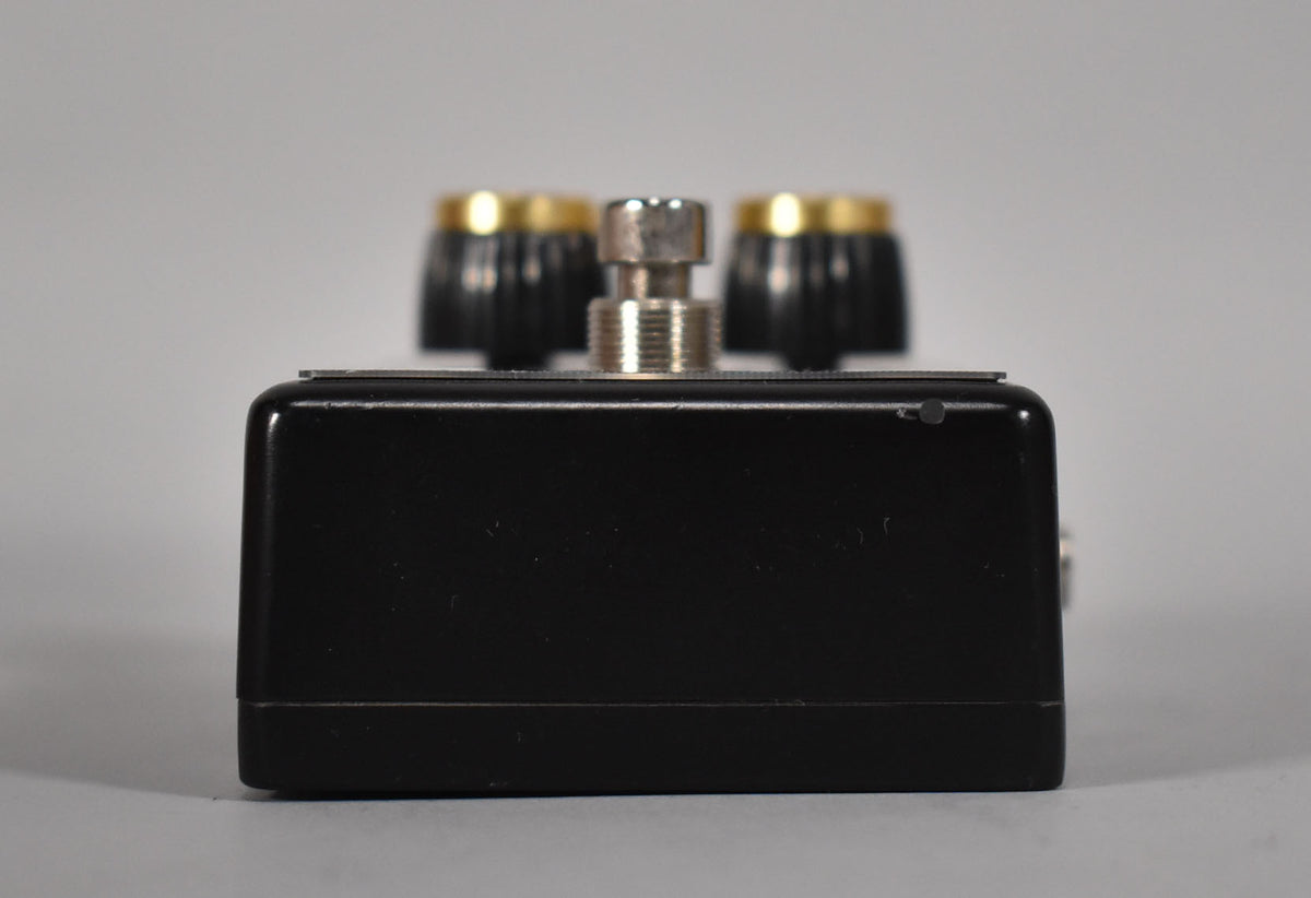 The black pedal is viewed from the side, showing a silver footswitch and two black and gold control knobs on top.
