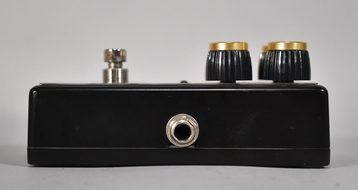 The black pedal is viewed from the side, showing a silver input jack, a footswitch, and two gold-topped black control knobs.