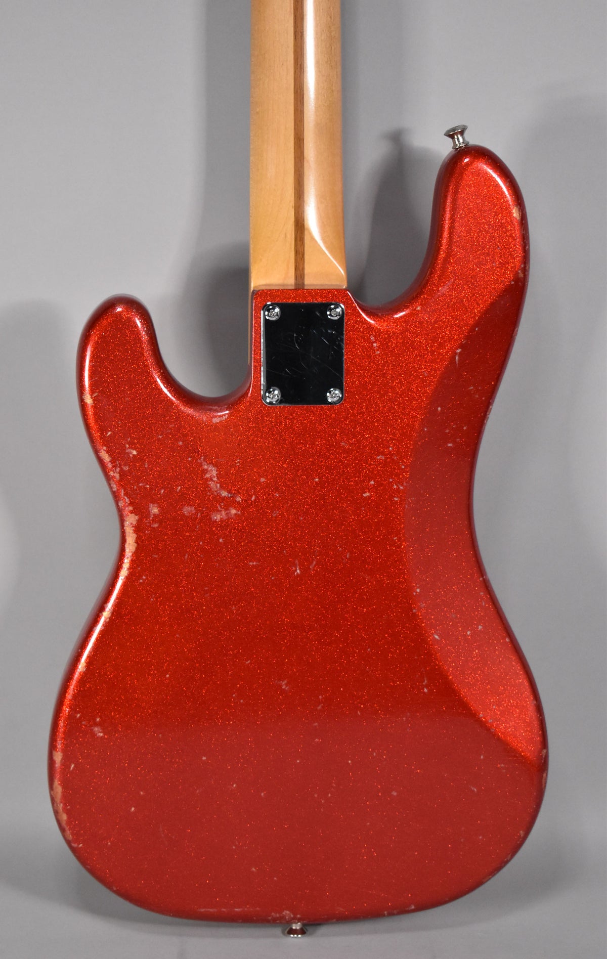 Rear view of a red sparkle P-style bass body, showing a maple neck, chrome neck plate, strap buttons, and heavy finish wear.