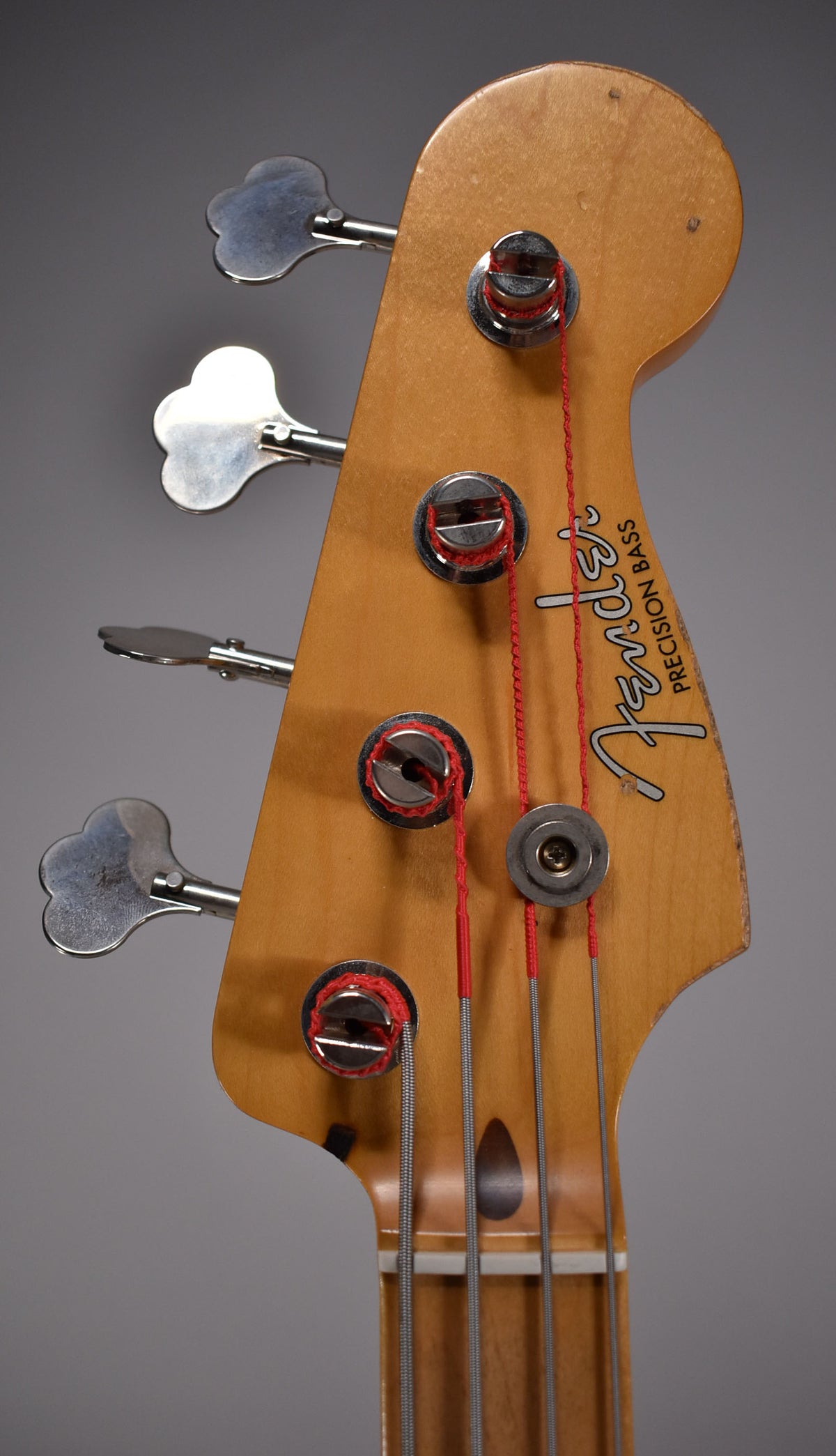 Close-up of the bass headstock, showing four chrome clover-leaf tuners, a string tree, and red-wrapped strings on a maple neck.