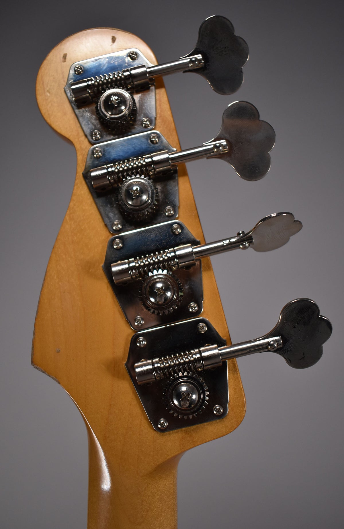 Close-up of the bass headstock from behind, showing four chrome clover-leaf tuners on a light wood neck with some wear.