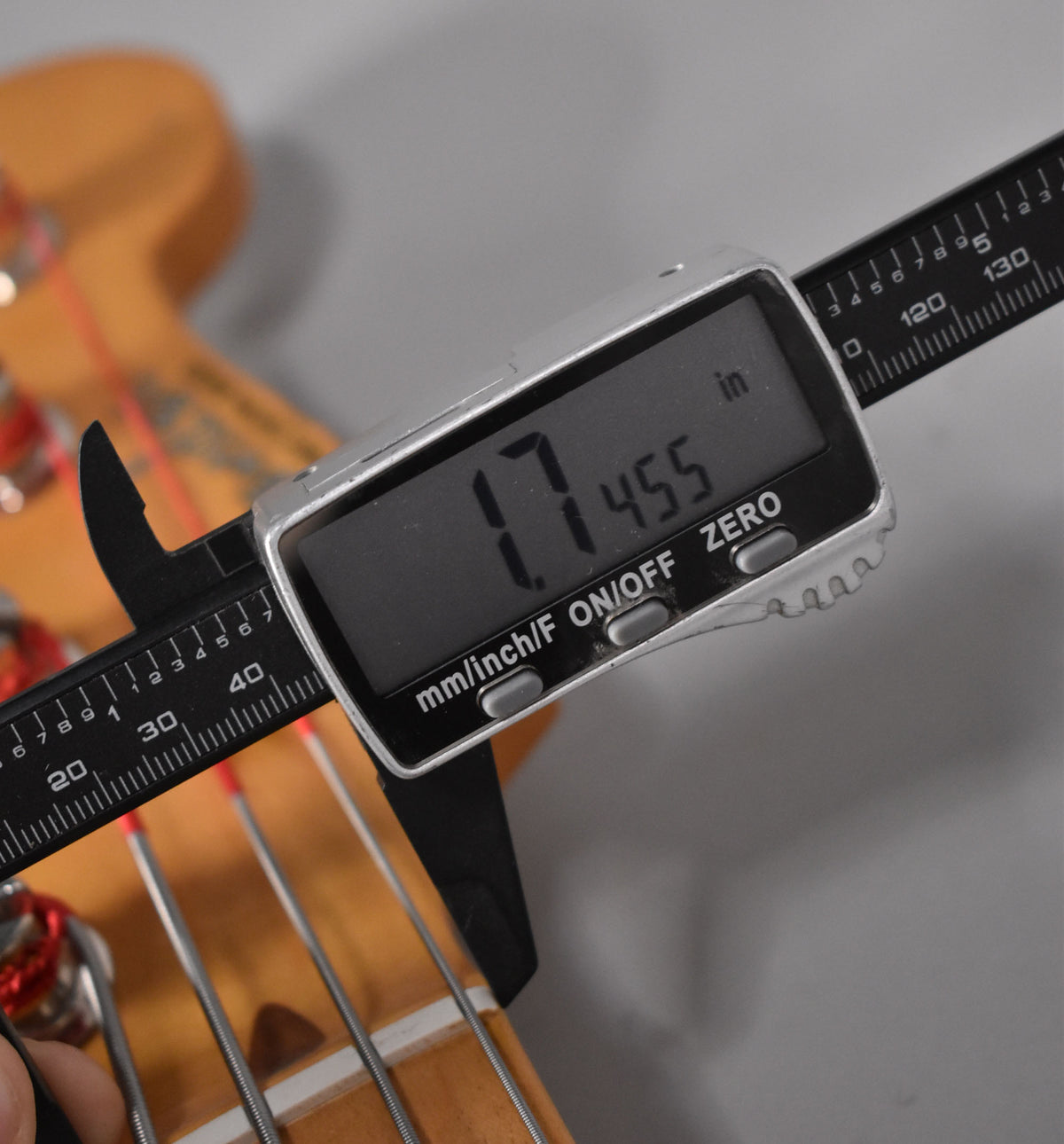 A digital caliper measures the bass's nut width at 1.7455 inches, with the maple neck and strings visible below.