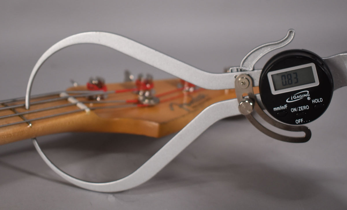 A digital caliper measures the bass's neck relief, showing 0.83mm, with the headstock and tuning pegs visible in the background.