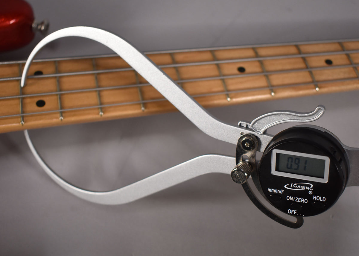 A digital caliper measures the bass's maple fretboard at the first fret, showing a reading of 0.91 inches.