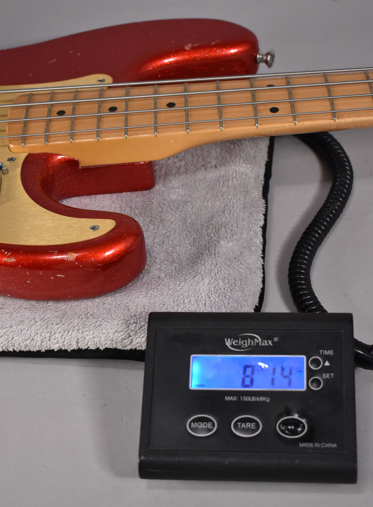 The red sparkle bass, with a gold pickguard and maple neck, rests on a scale displaying 8 lbs 14 oz.