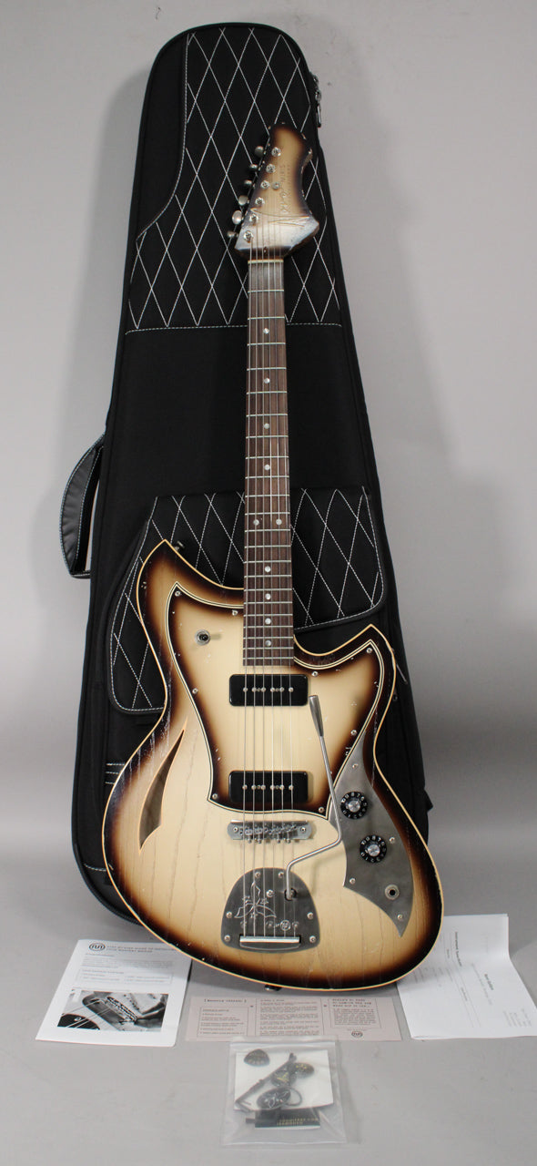 Full-length view of a light-aged semi-hollow electric guitar with a toasted marshmallow finish, two P90 pickups, and a vibrato tailpiece, standing in a black gig bag.