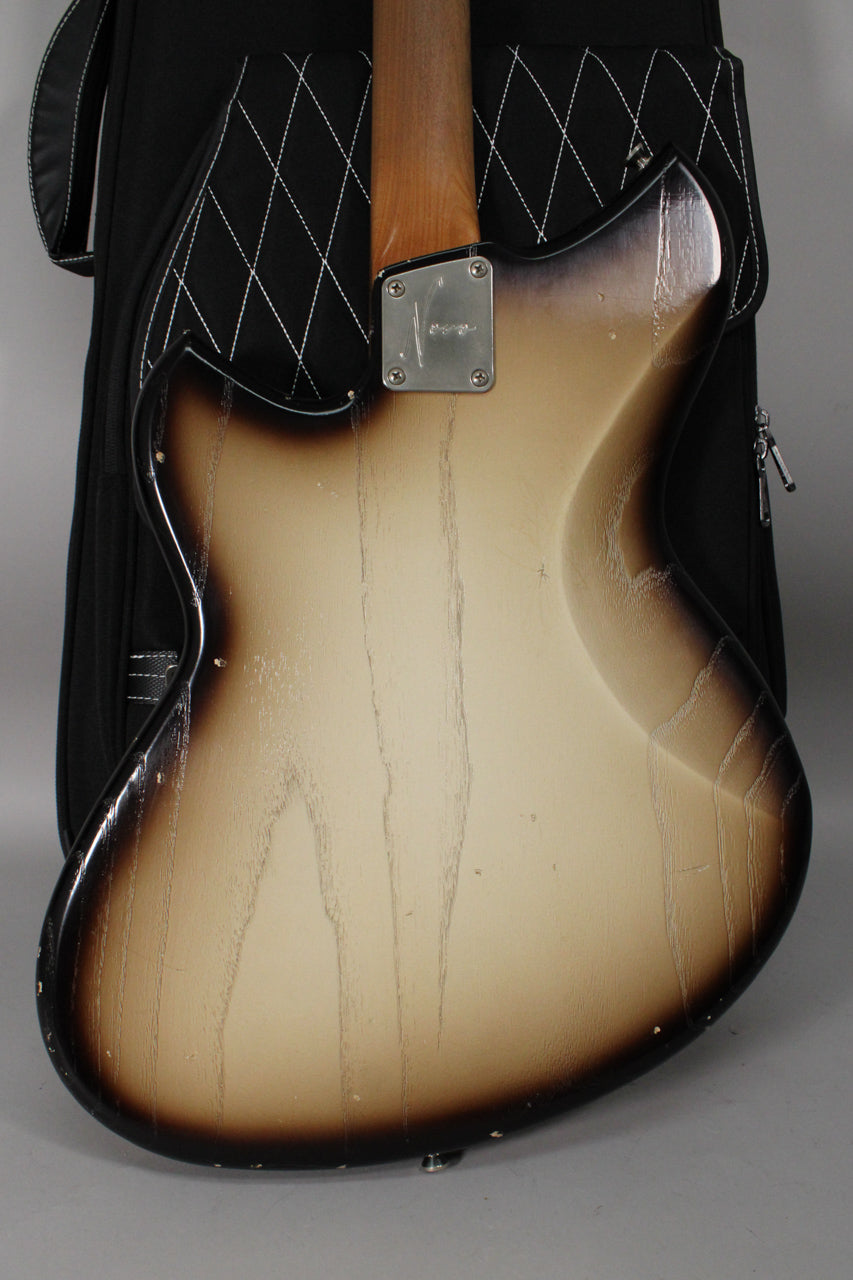 Back of a semi-hollow guitar with a light aged toasted marshmallow finish, showing a neck plate and a black gig bag.