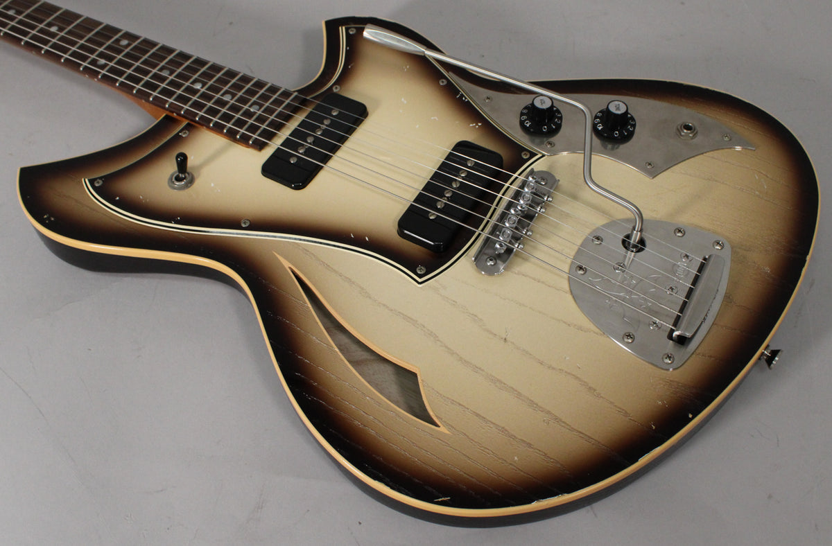The semi-hollow guitar's body, with f-hole, has a light aged toasted marshmallow finish, two black pickups, and a vibrato tailpiece.