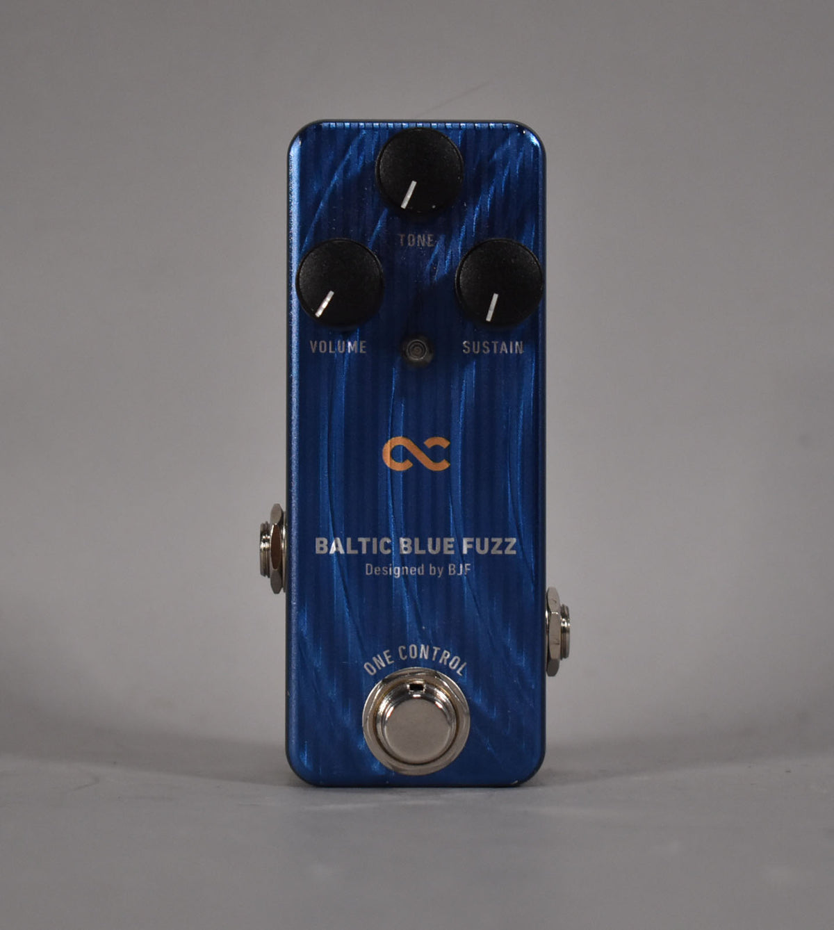 A top-down view of a metallic blue fuzz pedal with three black control knobs for Tone, Volume, and Sustain, and a silver footswitch.