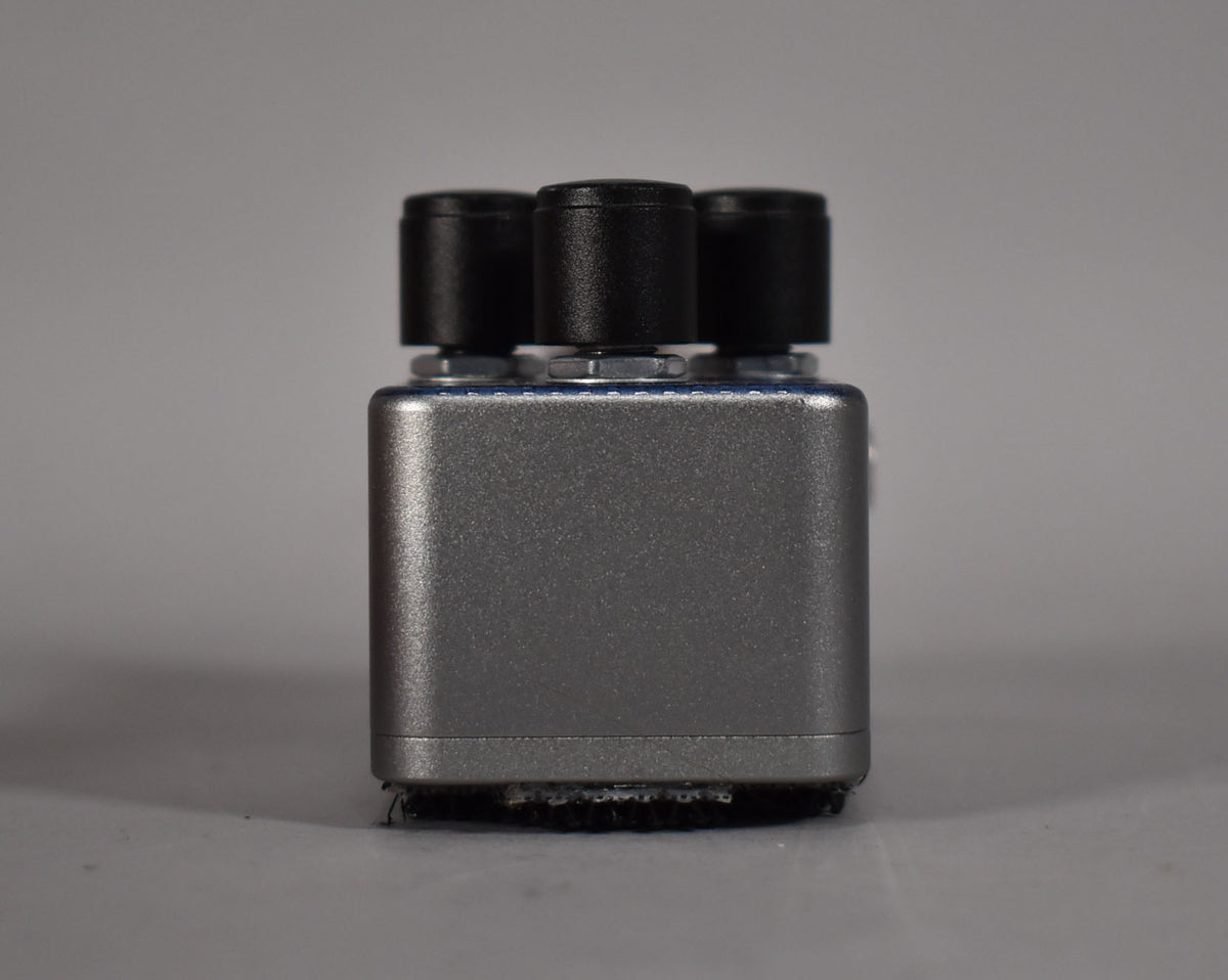 The pedal is viewed from the side, showing its textured silver casing, three black control knobs, and black velcro on the base.