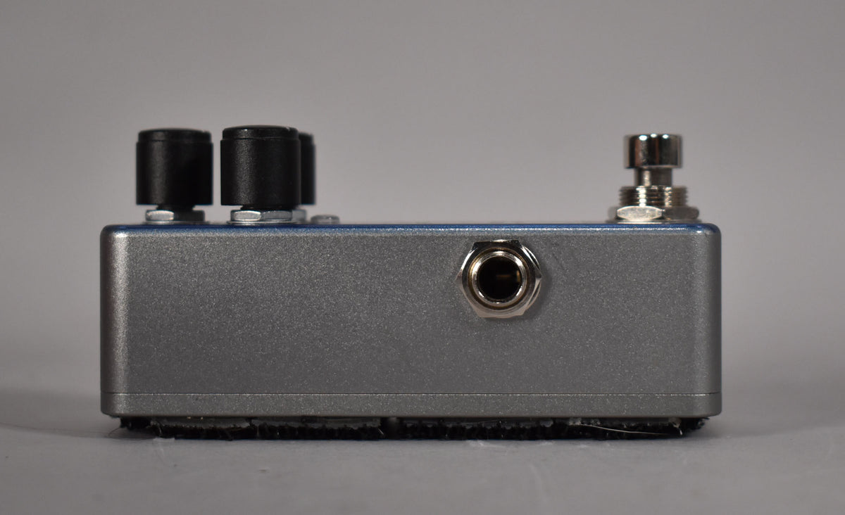Side view of a blue-gray pedal with two black knobs, a silver input jack, a silver footswitch, and black velcro on the bottom.