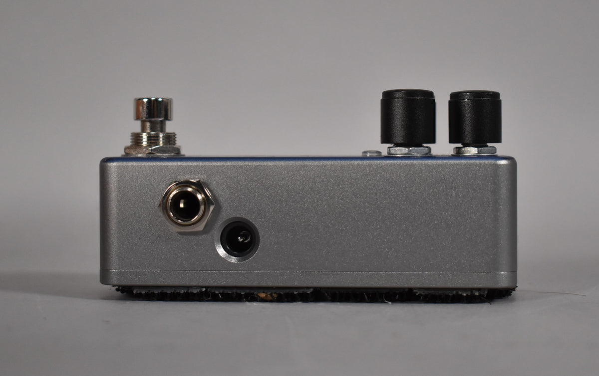 Side view of a gray pedal with a chrome input jack, power input, and two black knobs.