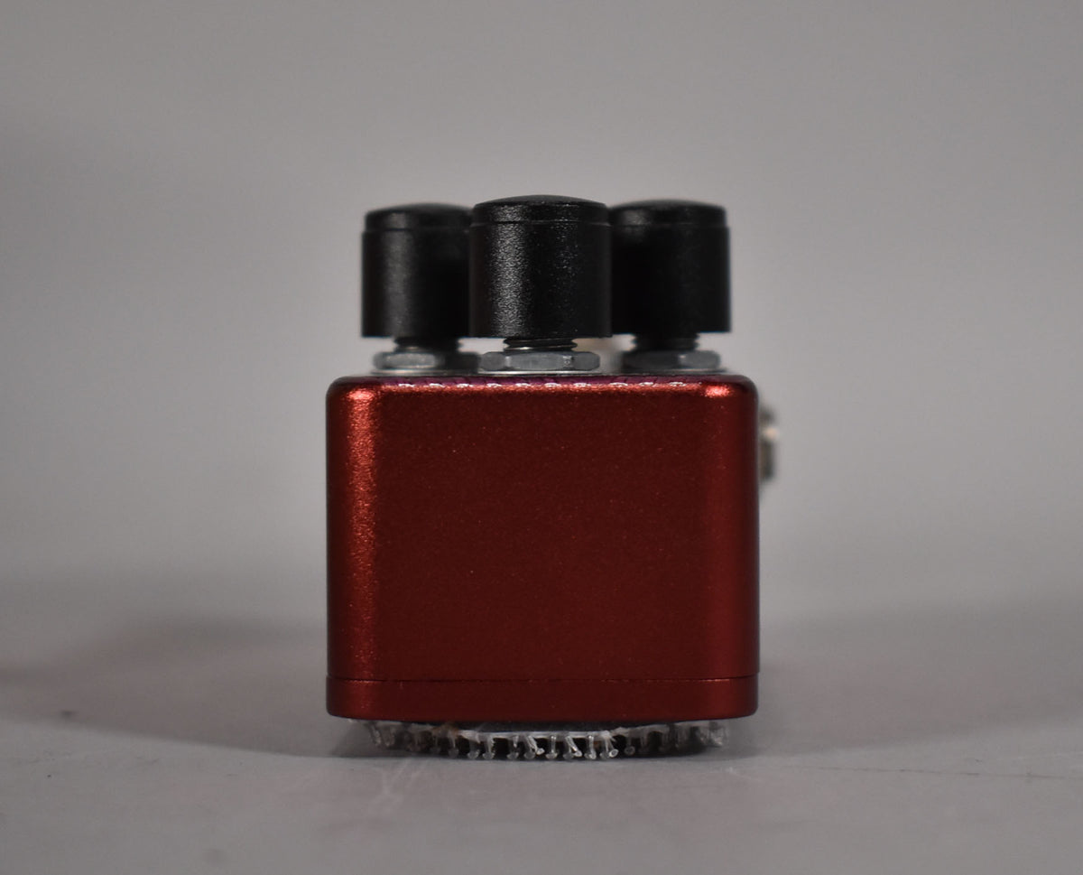 The cranberry red pedal is seen from the side, revealing three black control knobs and a textured silver bottom plate.